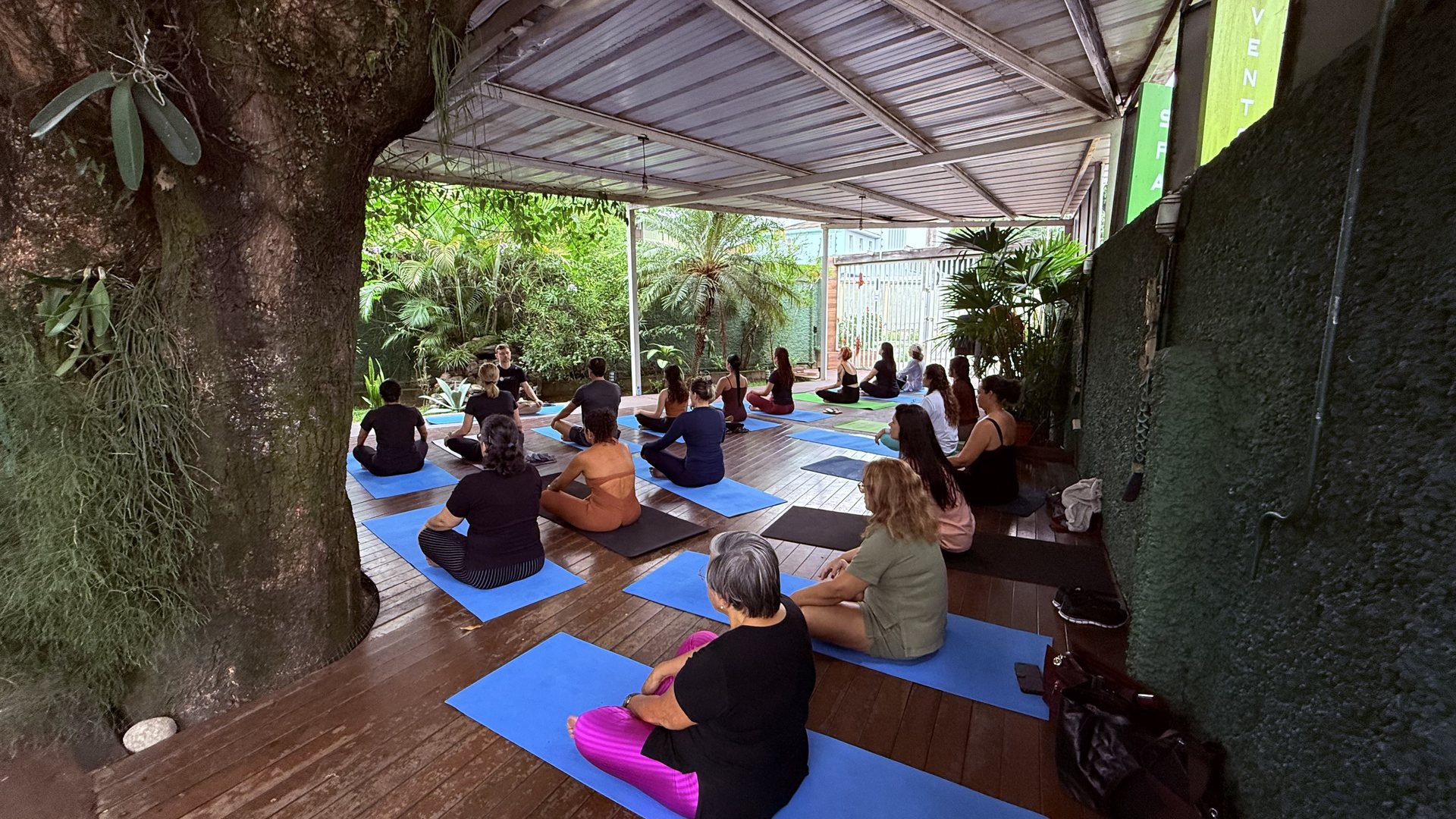 Aula de Yoga do Marzen no deck coberto, com alunos praticando em tapetes sob luz natural entre árvores e plantas